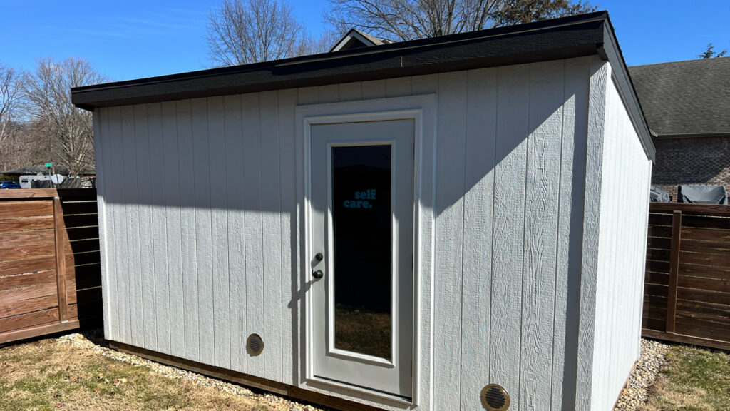 Gray outdoor sauna shed with a glass-panel door in a fenced backyard, built by Sauna Craft