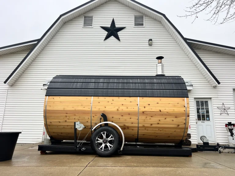 Finished custom trailer sauna built by Sauna Craft, side view on its trailer: cedar staves, steel bands, dark roof and wood-stove chimney